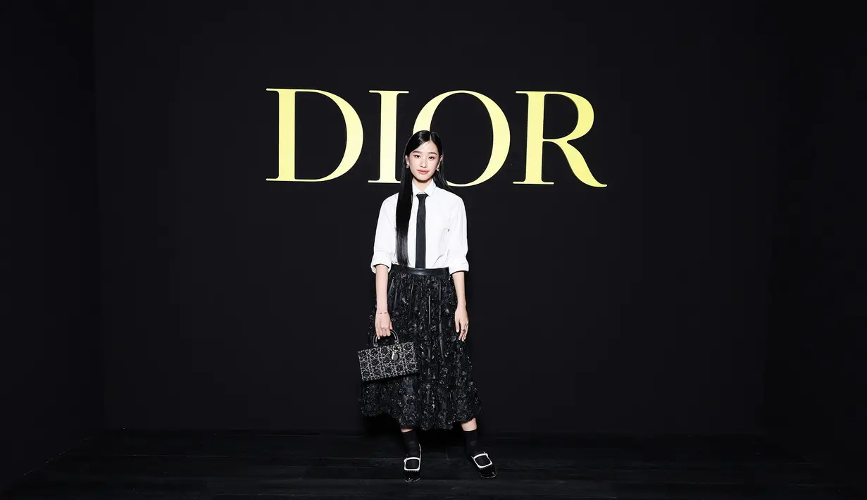 Tontawan Tantivejakul mengenakan kemeja katun putih Dior Winter 2023-2024 dengan rok kulit bordir. Dia juga mengenakan tas Dior, dasi hitam, kaus kaki, dan sepatu. [Dior]