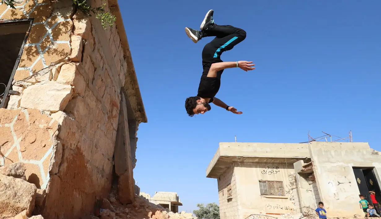 Warga menyaksikan pemuda Suriah melakukan parkour di atas reruntuhan bangunan yang hancur akibat serangan pasukan rezim di kota Binnish, Idlib pada 17 Juni 2020. Parkour merupakan olahraga ketangkasan yang terdiri dari lari, melompat, salto dan berayun. (Abdulaziz KETAZ / AFP)