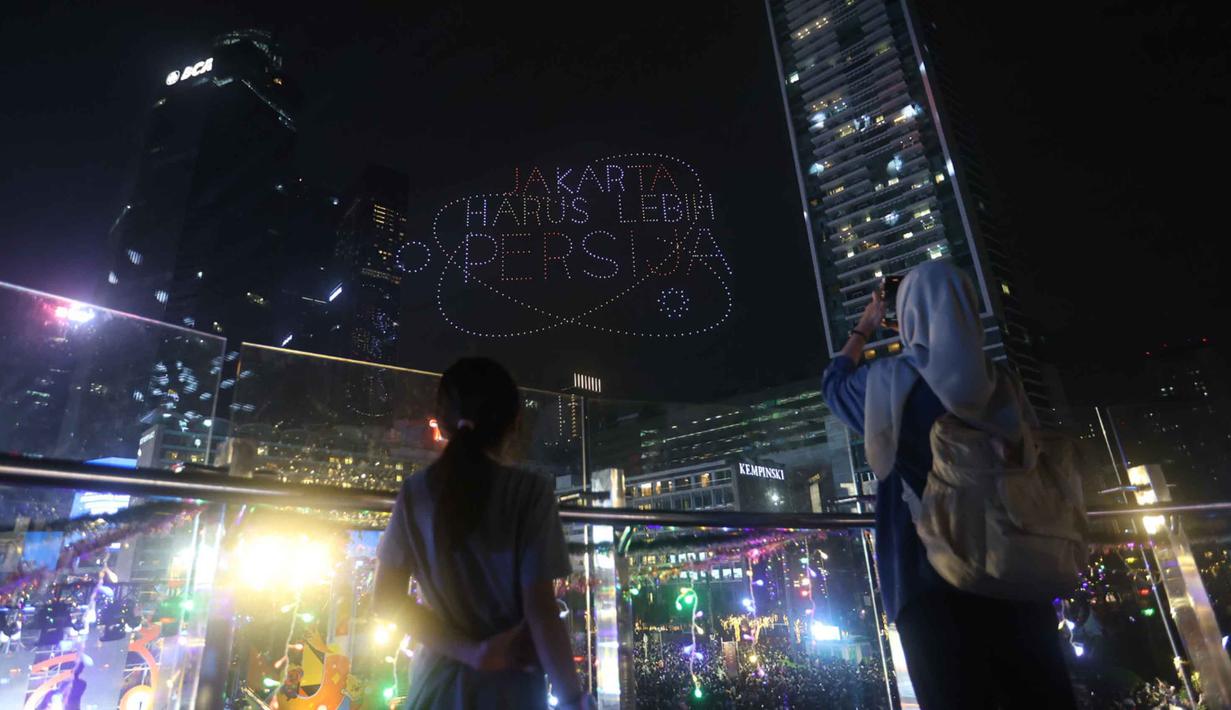 Dan ketika, visual drone membentuk tulisan “Jakarta Harus Lebih Persija”, warga antusias memberikan tepuk tangan dan sorakan meriah. Tampak dalam foto, warga mengabadikan pertunjukan drone saat perayaan malam pergantian tahun di kawasan Bundaran Hotel Indonesia, Jakarta, Rabu (31/12/2025). (Kapanlagi.com/Budy Santoso)