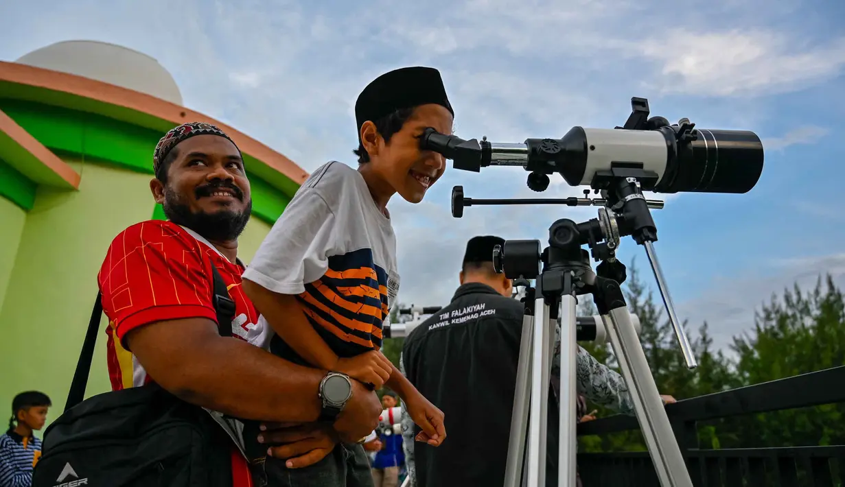 Untuk diketahui, metode rukyat sangat berbeda dengan hisab. Hisab murni menggunakan perhitungan matematis-astronomis tanpa memerlukan verifikasi visual di lapangan. Tampak dalam foto, seorang pria menggendong anaknya untuk mengamati bulan, menjelang Idul Fitri sekaligus menandai berakhirnya bulan suci Ramadan1447 Hijriah melalui teleskop di kantor observatorium, pantai Lhoknga, Aceh pada Kamis 19 Maret 2026. (CHAIDEER MAHYUDDIN/AFP)