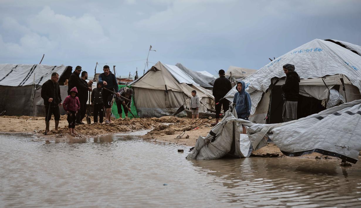 Hujan deras dan angin kencang merendam tenda-tenda para pengungsi, dan memperburuk krisis kemanusiaan yang sudah ada. Tampak dalam foto, warga Palestina berjalan melewati area yang tergenang banjir di kamp tenda sementara setelah hujan lebat di Kota Gaza, Kamis, 26 Maret 2026 (AP Photo/Jehad Alshrafi)