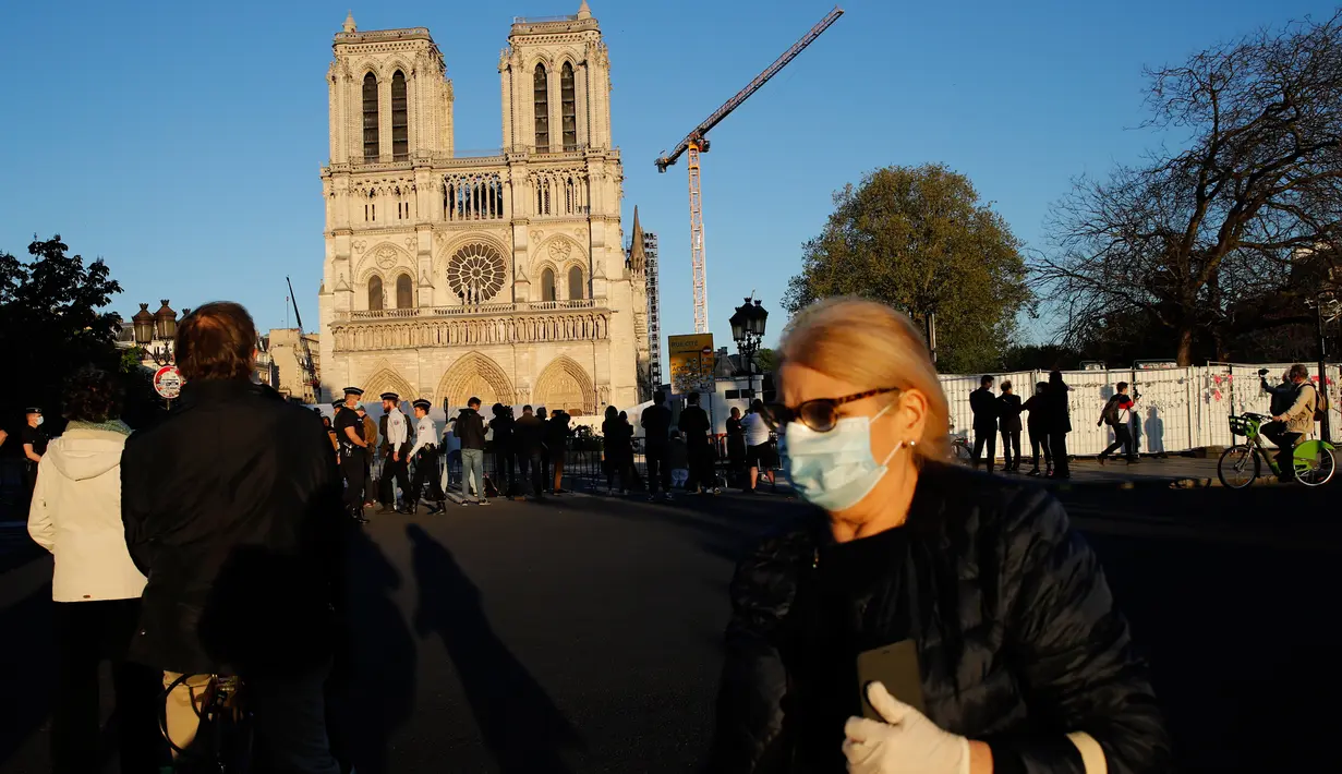 Seorang perempuan melintasi orang-orang yang berhenti di depan Katedral Notre Dame saat kembali membunyikan lonceng utamanya di Paris, Rabu (15/4/2020). Lonceng utama katedral kembali dibunyikan setelah satu tahun mengalami kebakaran yang sampai mengejutkan seluruh warga dunia. (AP/Christophe Ena)