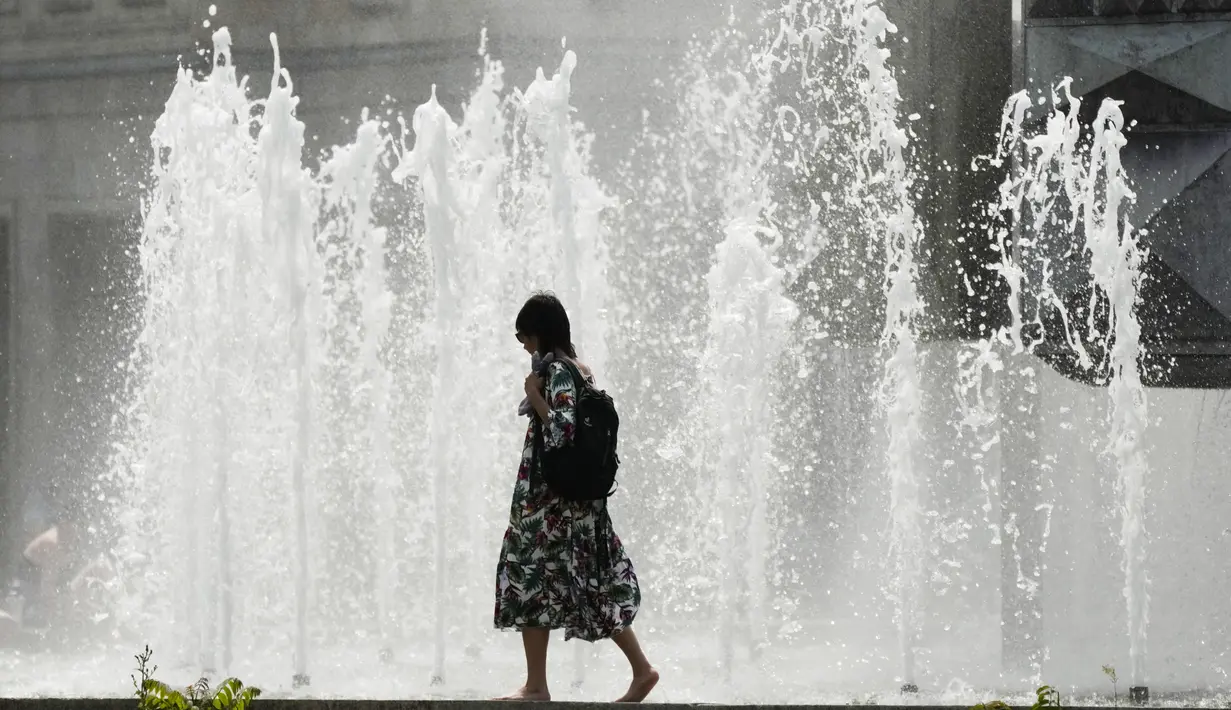 Seorang perempuan berjalan melewati air mancur pada hari musim panas di Berlin, Minggu (19/6/2022). Di Jerman, di mana suhu tertinggi diperkirakan 38 derajat celcius (100,4 F), menteri kesehatan mendesak kelompok rentan menjaga tubuh tetap terhidrasi. (AP Photo/Markus Schreiber)