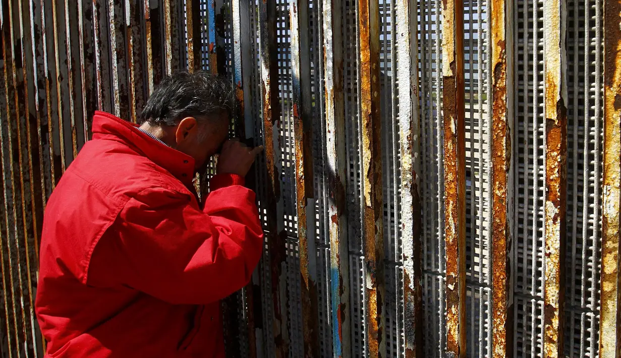 Seorang warga Meksiko terbujur kaku sambil memegang pagar perbatasan sambil berbincang dengan keluarganya di Playas Tijuana, Meksiko, (12/4). Tembok ini memisahkan antara Meksiko dan Amerika Serikat. (REUTERS / Jorge Duenes)