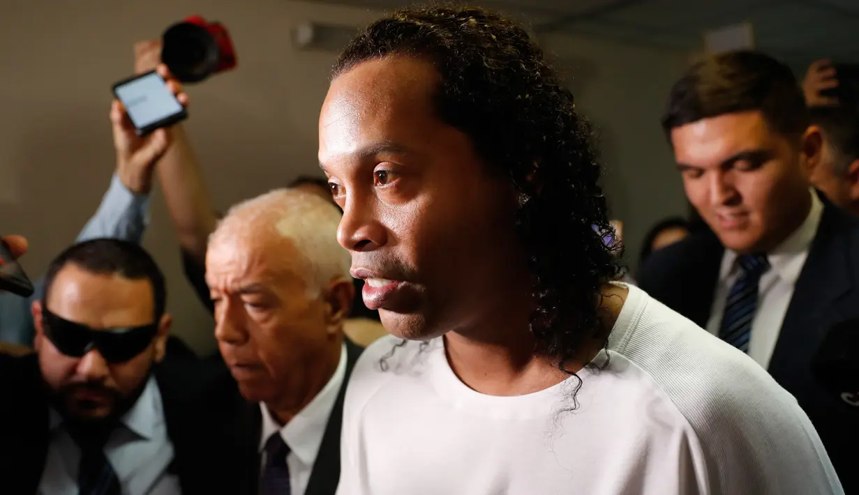 Mantan pemain timnas Brasil, Ronaldinho dikawal oleh polisi meninggalkan kantor pengadilan di Asuncion, ibu kota Paraguay, Jumat (6/3/2020). Ronaldinho bersama saudara laki-lakinya berurusan dengan pihak berwenang Paraguay karena menggunakan paspor palsu untuk memasuki negara itu. (AP/Jorge Saenz)