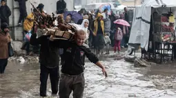 Laporan media internasional menyebutkan cuaca dingin memperberat penderitaan anak-anak dan para lanjut usia di tengah keterbatasan selimut, pakaian musim dingin, serta alat pemanas. Tampak dalam foto, orang-orang membawa kayu sambil berjalan di tengah banjir di kamp darurat yang menampung warga Palestina yang terlantar setelah hujan deras di Kota Gaza pada Selasa 25 November 2025. (Omar AL-QATTAA/AFP)