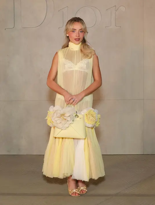 Sabrina Carpenter tampil mengenakan long dress butter yellow berbahan tulle transparan kerah tinggi. Dress sleeveless tersebut semakin menarik dengan aksen bunga 3D besar di bagian rok. Melengkapi tampilannya dengan membawa tas Dior warna senada. [Dok. DIOR]