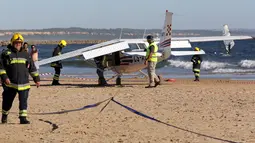 Petugas pemadam berdiri di samping sebuah pesawat kecil yang mendarat darurat di pantai Sao Joao yang ramai dekat Lisbon, Portugal, Rabu (2/8). Pesawat kecil yang diketahui jenis cessna itu membawa dua orang termasuk pilot. (AP Photo/Armando Franca)