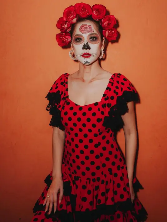 Wajahnya dirias dengan makeup tengkorak tradisional yang dikenal sebagai “calavera,” lengkap dengan detail rumit dan bunga mawar pada dahinya, mencerminkan penghormatan kepada roh leluhur dalam perayaan ini. [@itsmebcl].