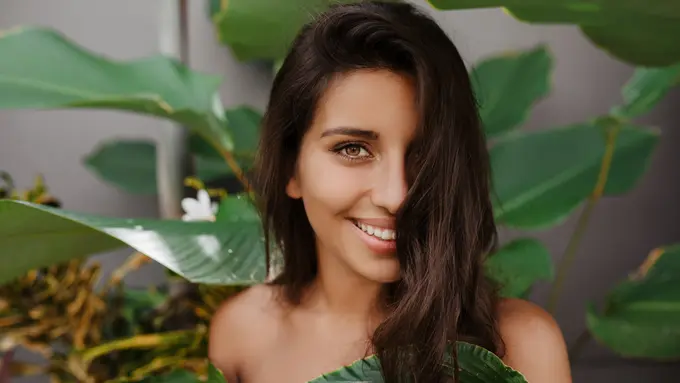 Seorang perempuan berdiri di antara daun dengan setengah wajah tertutup oleh rambut (Foto Dok: Freepik/lookstudio)