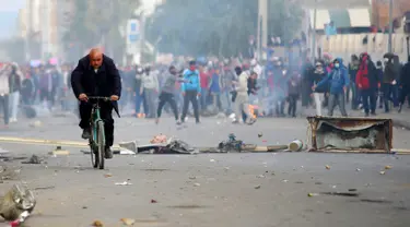 Seorang pria mengendarai sepeda saat pengunjuk rasa melemparkan batu ke arah polisi di luar kantor pemerintah setempat di Kasserine, Tunisia, Kamis (21/1). Mereka menuntut solusi dari masalah pengangguran yang meliputi negaranya. (REUTERS/Amine Ben Aziza)