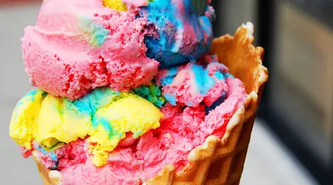 Es krim pelangi dengan lapisan warna merah, kuning, hijau, biru, dan ungu, disajikan dalam cone dengan topping sprinkle ceria