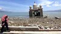 Warga melintas di depan Masjid Terapung Arqam Bab Al Rahman pasca gempa dan tsunami Palu di Pantai Talise, Sulawesi Tengah. Bangunan masjid yang terletak di pinggir pantai terlihat utuh meski sebagian bangunannya tenggelam. (Liputan6.com/Fery Padolo)
