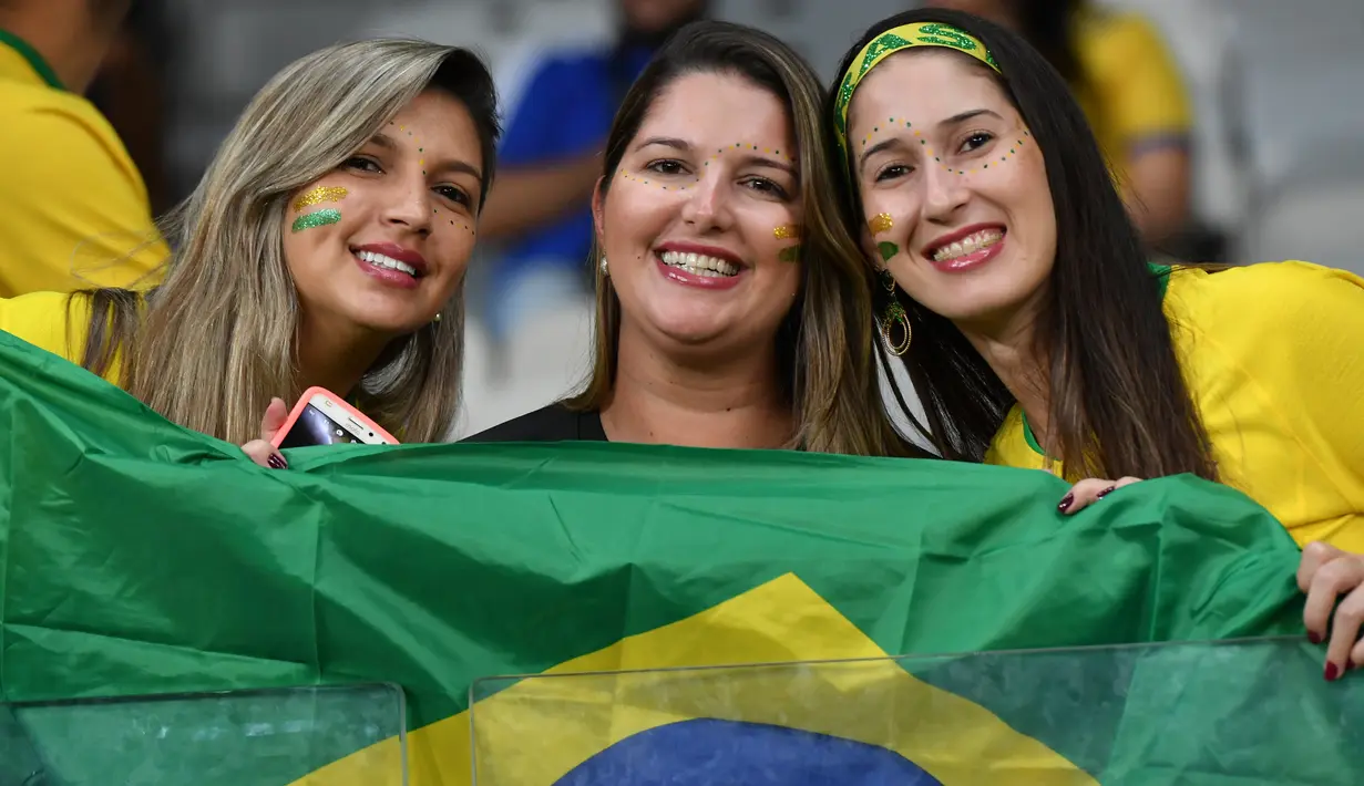 Tiga fans wanita tersenyum sambil memegang bendera Brasil sebelum pertandingan melawan Argentina pada semifinal Copa America 2019 di Stadion Mineirao (2/7/2019). Di pertandingan ini Brasil menang 2-0 atas Argentina berkat gol striker Gabriel Jesus dan Roberto Firmino. (AFP Photo/Nelson Almeida)