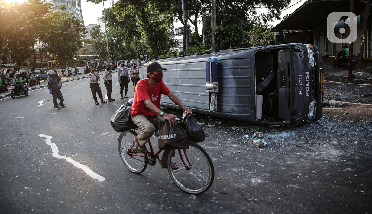 Seorang pengendara sepeda melintas dekat sebuah mobil polisi yang dirusak massa saat bentrok di kawasan Pejompongan, Jakarta, Rabu (7/10/2020). (Liputan6.com/Faizal Fanani)
