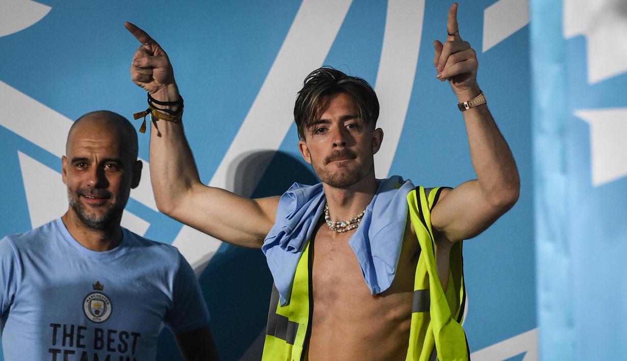 Pemain Manchester City, Jack Grealish (kanan), memasuki panggung dengan mengenakan rompi proyek setelah melakukan parade Treble Winners, Selasa (13/6/2023) dini hari WIB. Pep Guardiola hanya bisa tersenyum melihat kelakuan anak asuhnya tersebut. (AFP/Oli Scarff)
