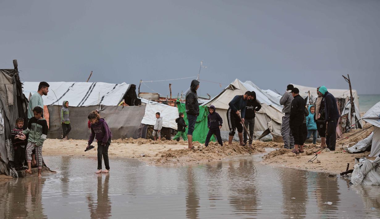 Para pengungsi terpaksa menggali parit secara manual di sekitar tenda untuk mengalirkan air. Tampak dalam foto, warga Palestina berjalan melewati area yang tergenang banjir di kamp tenda sementara setelah hujan lebat di Kota Gaza, Kamis, 26 Maret 2026 (AP Photo/Jehad Alshrafi)