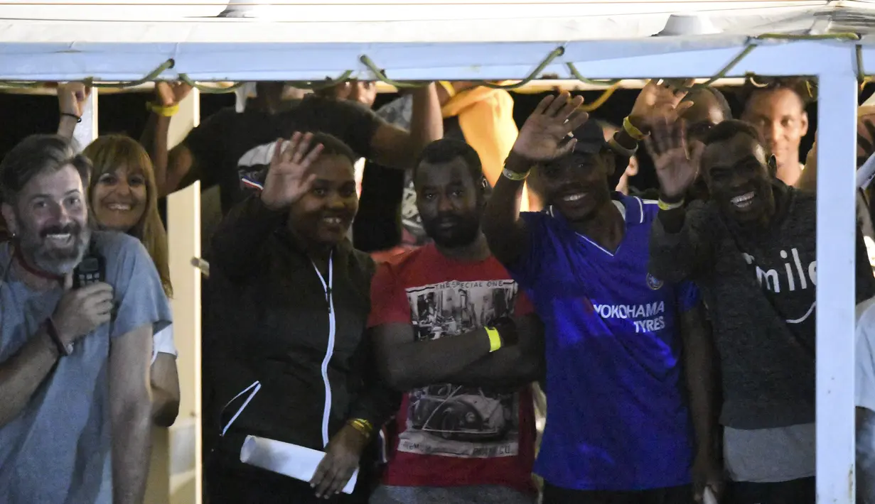 Para imigran melambaikan tangan di kapal Open Arms sebelum turun di pulau Sisilia Lampedusa, Italia (20/8/2019). Jaksa penuntut Italia Luigi Patronaggio memerintahkan penyitaan sebuah kapal penyelamat migran dan evakuasi langsung dari 80 migran yang tersisa di kapal. (AP Photo/Salvatore Cavalli)