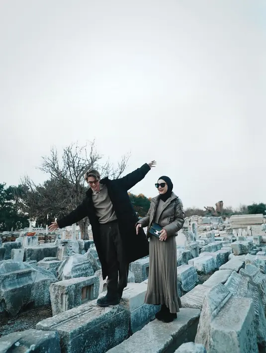Pasutri Rey Mbayang dan Dinda Hauw tampil serasi dengan outfit berwarna cokelat. Rey Mbayang mengenakan polo shirt dan celana panjang cokelat gelap yang serasi, ditumpuknya dengan long coat hitam. Sedangkan Dinda Hauw tampil dengan inner cokelat yang serasi dengan knitted skirtnya, ditumpuk dengan puffer jacket yang serasi, dan hijab hitam polos. [Foto: Instagram/rey_mbayang]