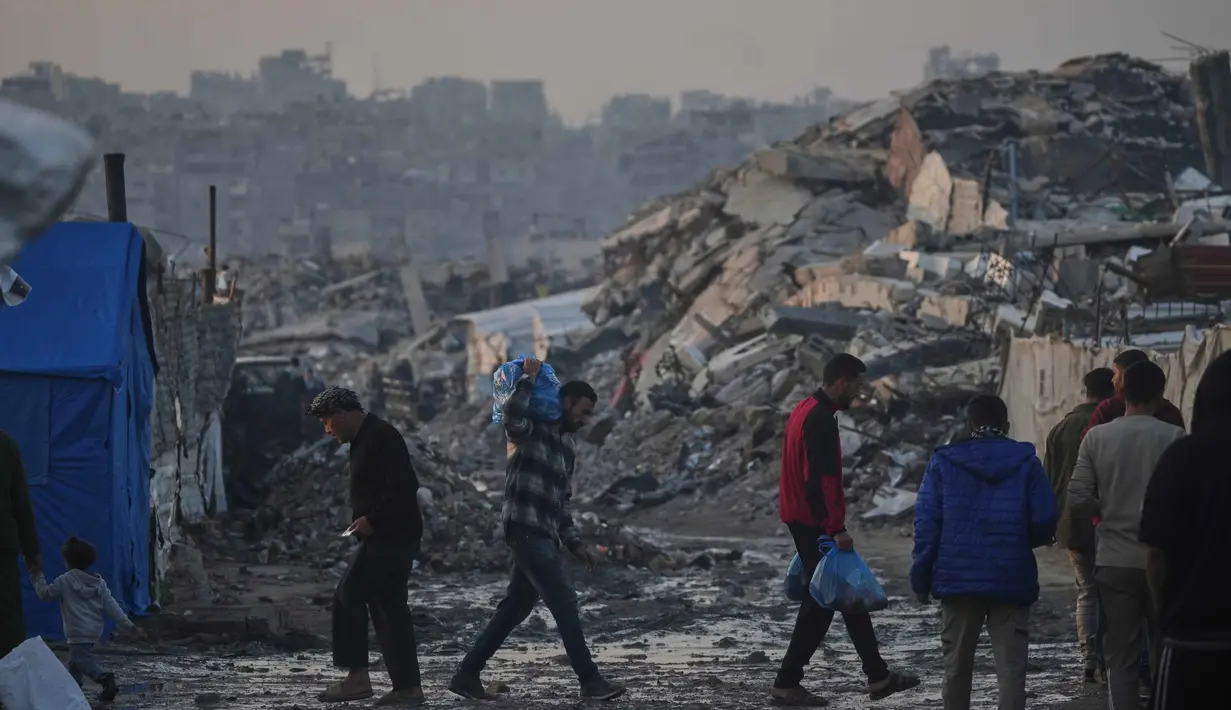 Fokus bantuan mencakup pengiriman pasokan medis ke fasilitas kesehatan yang tersisa serta peralatan untuk perbaikan layanan air dan sanitasi guna mencegah krisis penyakit. Tampak dalam foto, warga Palestina berjalan melewati perkemahan tenda di tengah bangunan-bangunan yang hancur akibat serangan udara dan darat Israel di Kota Gaza, Selasa 6 Januari 2026. (AP Photo/Jehad Alshrafi)