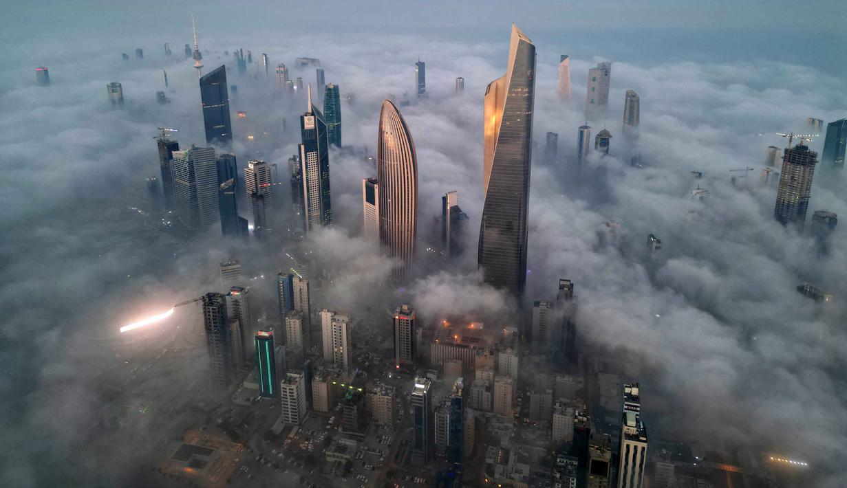 Secara umum, di Kuwait, kabut tebal lebih sering muncul selama musim dingin (Desember hingga Maret) karena suhu malam hari yang lebih rendah dan kelembapan yang lebih tinggi. Tampak foto udara memperlihatkan gedung pencakar langit yang tertutup kabut pagi di Kota Kuwait pada Minggu 9 November 2025. (YASSER AL-ZAYYAT/AFP)
