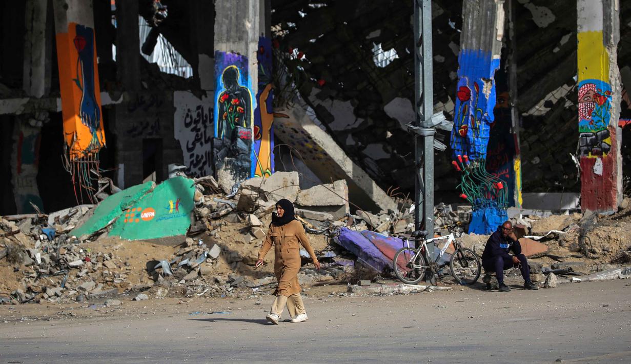 Diketahui, Israel masih menduduki lebih dari 50 persen wilayah Gaza meski ada perjanjian gencatan senjata. Tampak dalam foto, seorang perempuan berjalan melewati lukisan-lukisan di sisa-sisa bangunan yang hancur di kamp pengungsi Maghazi di luar Deir al-Balah pada Senin 8 Desember 2025. (EYAD BABA/AFP)