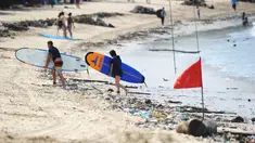 Turis berjalan dengan papan selancar melintasi puing-puing dan sampah di pesisir Pantai Kuta, Bali, Minggu (9/12). Kawasan pantai Kuta kembali dipenuhi oleh sampah hanyut terbawa oleh gelombang. (SONNY TUMBELAKA / AFP)
