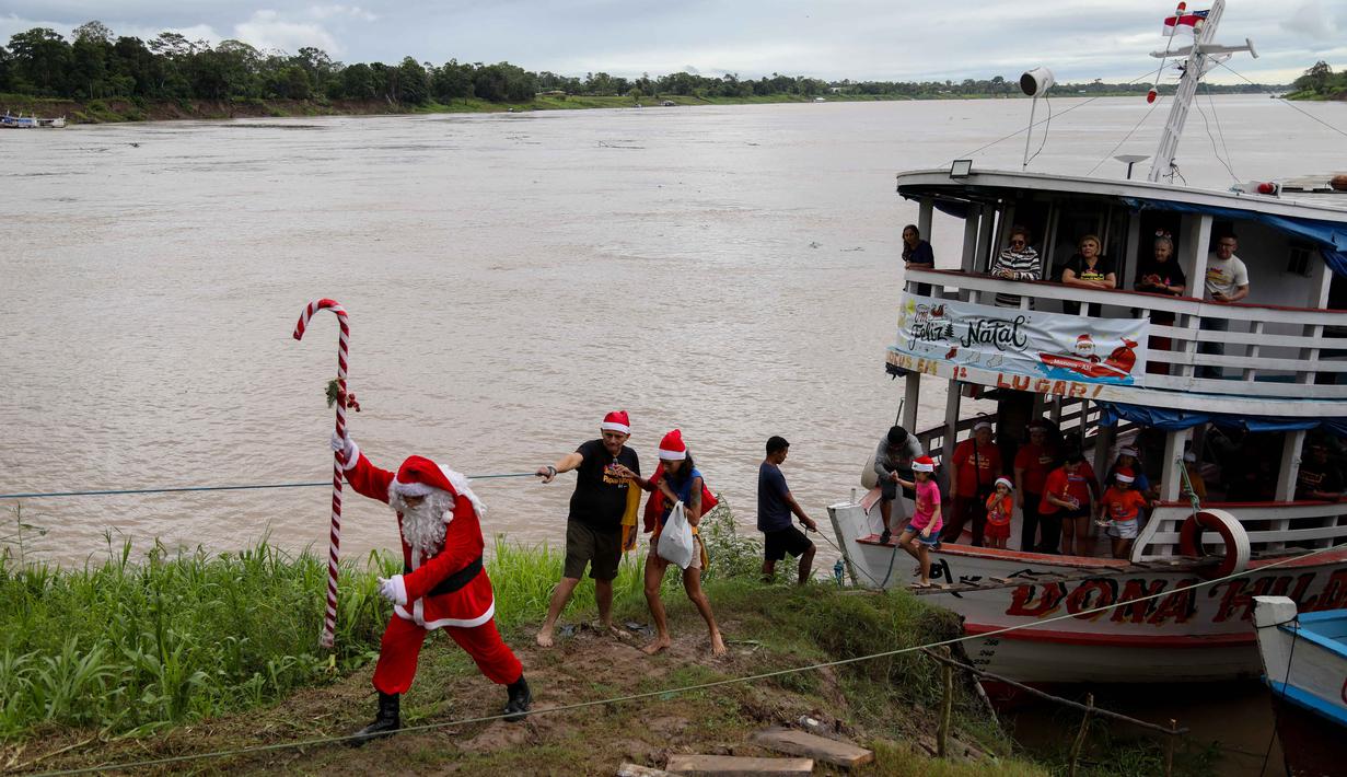 Untuk informasi, sudah sejak tahun 1998 kelompok relawan Sahabat Sinterklas ini berbagi kebahagiaan Natal untuk anak-anak di komunitas pedesaan dan tepi sungai di hutan hujan Amazon. Tampak dalam foto, relawan Jorge Alberto Barrozo, yang mengenakan kostum Sinterklas, berjalan untuk mengantarkan hadiah kepada anak-anak di komunitas tepi sungai di daerah pedesaan Careiro da Varzea, negara bagian Amazonas, Brasil pada Sabtu 20 Desember 2025. (Michael DANTAS/AFP)