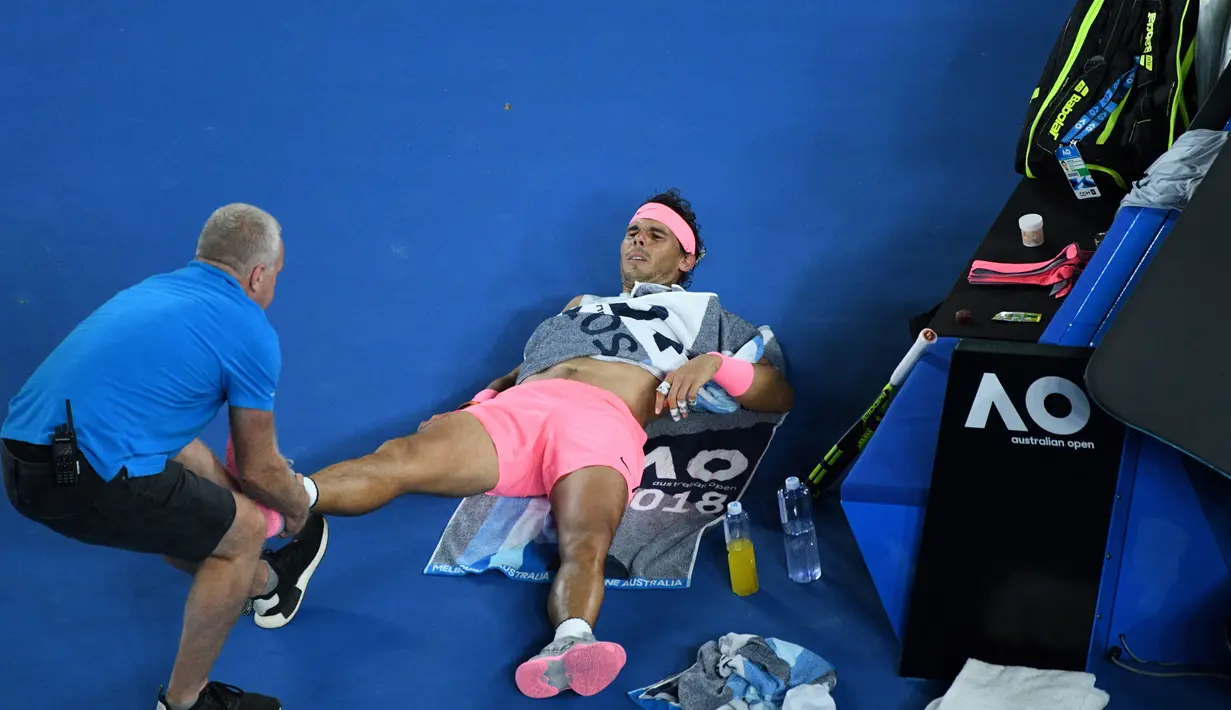 Petenis Spanyol, Rafael Nadal mendapat perawatan dari pelatihnya terkait cedera paha kanan pada perempat final Australia Terbuka 2018 di Melbourne, Selasa (23/1). Nadal mundur di set kelima saat menghadapi petenis Kroasia, Marin Cilic. (WILLIAM WEST/AFP)