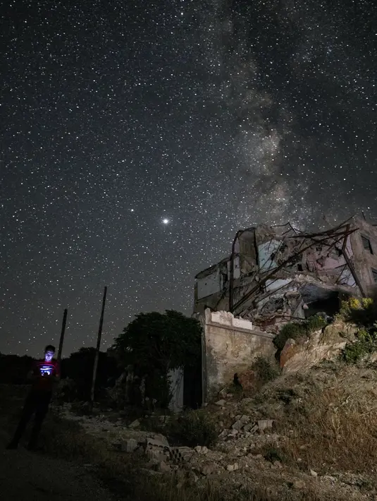 Galaksi Bima Sakti menghiasi langit malam saat seorang pria menggunakan ponselnya dekat gedung-gedung yang hancur akibat pengeboman di Kota Ariha, Provinsi Idlib, Suriah, Sabtu (27/6/2020). (Omar HAJ KADOUR/AFP)