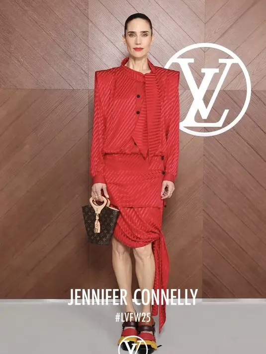 Jennifer Connelly tampil serba merah mengenakan atasan lengan panjang dengan kerah pita dipadukan mini skirt berpita samping. Sambil membawa bucket bag LB coklat dan heels strap warna-warni. [dok. Louis Vuitton]