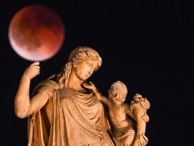 Foto ini menampilkan bulan purnama yang juga dikenal sebagai “Blood Moon” selama gerhana bulan, dengan patung dewi Yunani kuno Irene yang memegang seorang anak di latar depan, di pusat Athena, Yunani pada 7 September 2025. (Angelos TZORTZINIS/AFP)