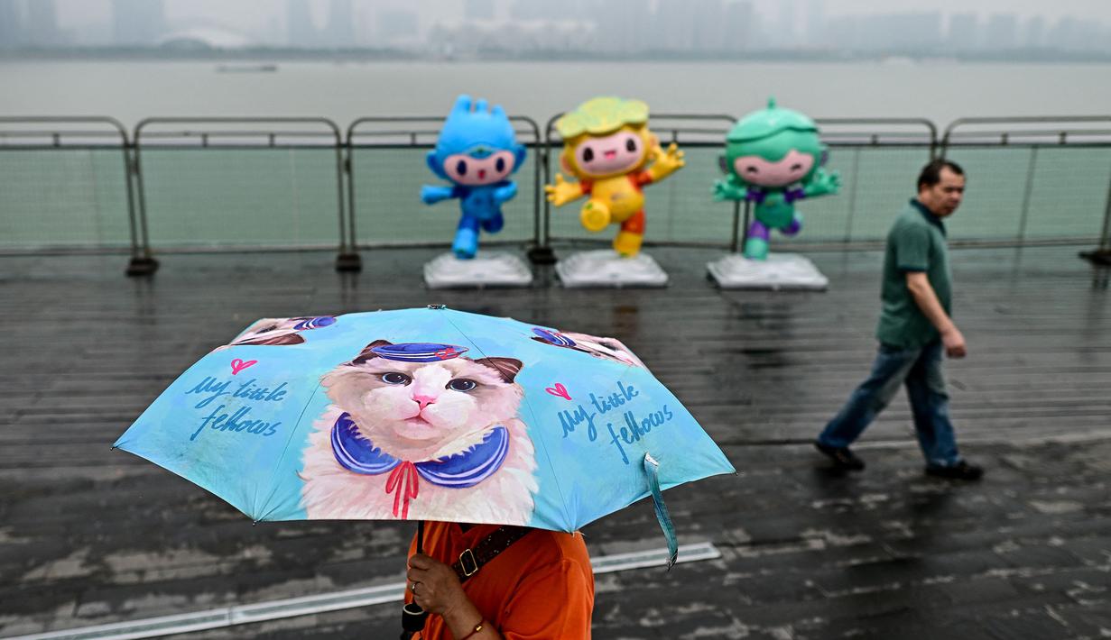 Seorang pejalan kaki menggunakan payung saat melintas di pedestrian Sungai Qiantang. Hangzhou, Zhejiang, China yang berhias maskot Asian Games 2022 (Chenchen, Congcong, dan Lianlian) pada 22 September 2023 waktu setempat. (AFP/Manan Vatsyayana)