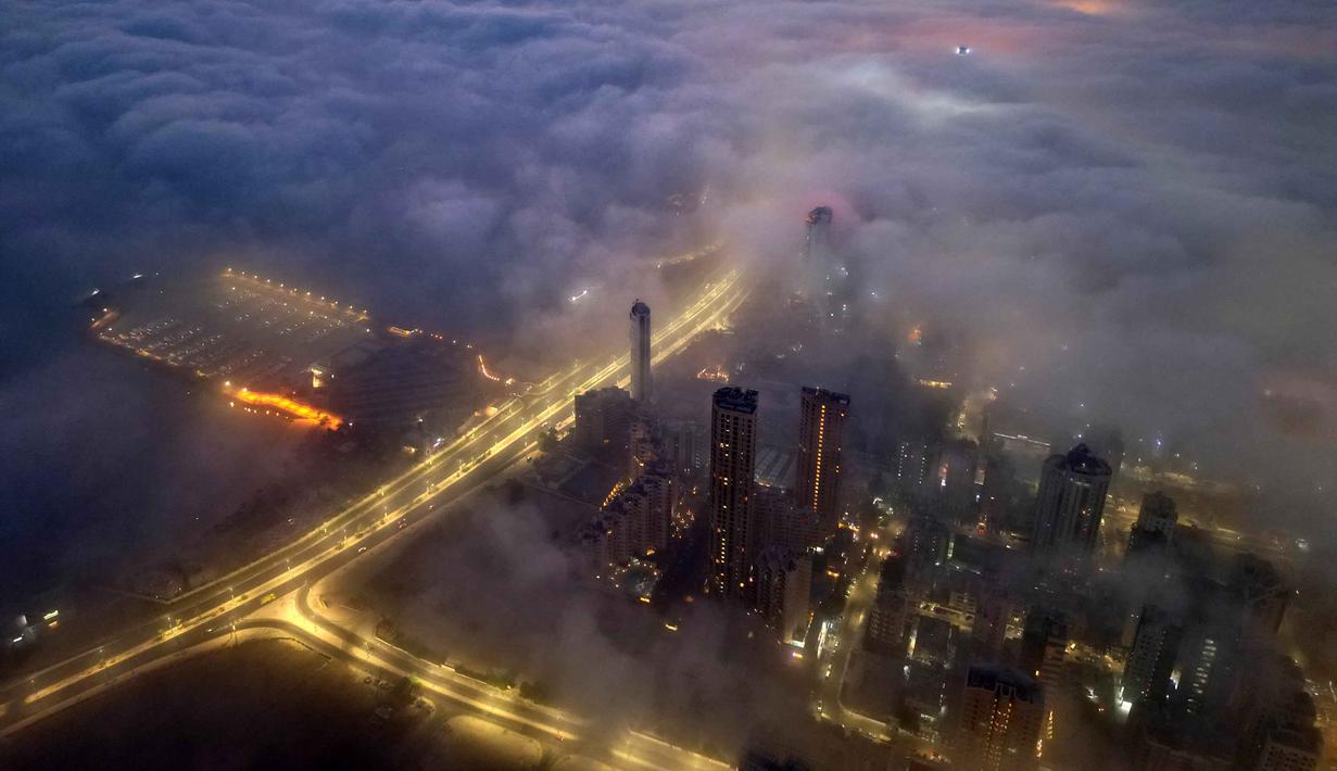 Kabut tebal sering terjadi di Kuwait selama musim dingin, yang menciptakan pemandangan gedung pencakar langit yang indah menembus lapisan awan rendah. Tampak foto udara memperlihatkan gedung pencakar langit dan jalan tol yang diterangi lampu, tertutup kabut pagi di Kota Kuwait pada Minggu 9 November 2025. (YASSER AL-ZAYYAT/AFP)