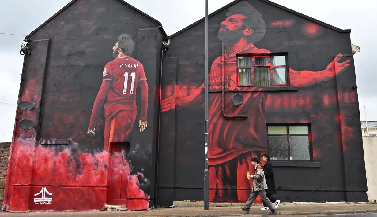 Dua pejalan kaki melewati mural seniman Inggris, John Culshaw yang menggambarkan pesepak bola Mesir Mohamed Salah di fasad bangunan dekat Stadion Anfield, di Liverpool, Rabu (30/3/2022). Salah sudah berada di urutan kesembilan dalam daftar pencetak gol sepanjang masa Liverpool. (Paul ELLIS / AFP)