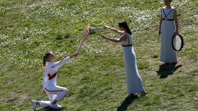 Aktris Yunani Xanthi Georgiou (tengah) sebagai Imam Besar menyalakan obor Olimpiade Tokyo 2020 yang dibawa juara menembak Olimpiade asal Yunani, Anna Korakaki (kiri) saat upacara penyalaan api Olimpiade Tokyo 2020 di Kota Tua Olympia, Yanani, Kamis (12/3/2020). (AP Photo/Yorgos Karahalis)