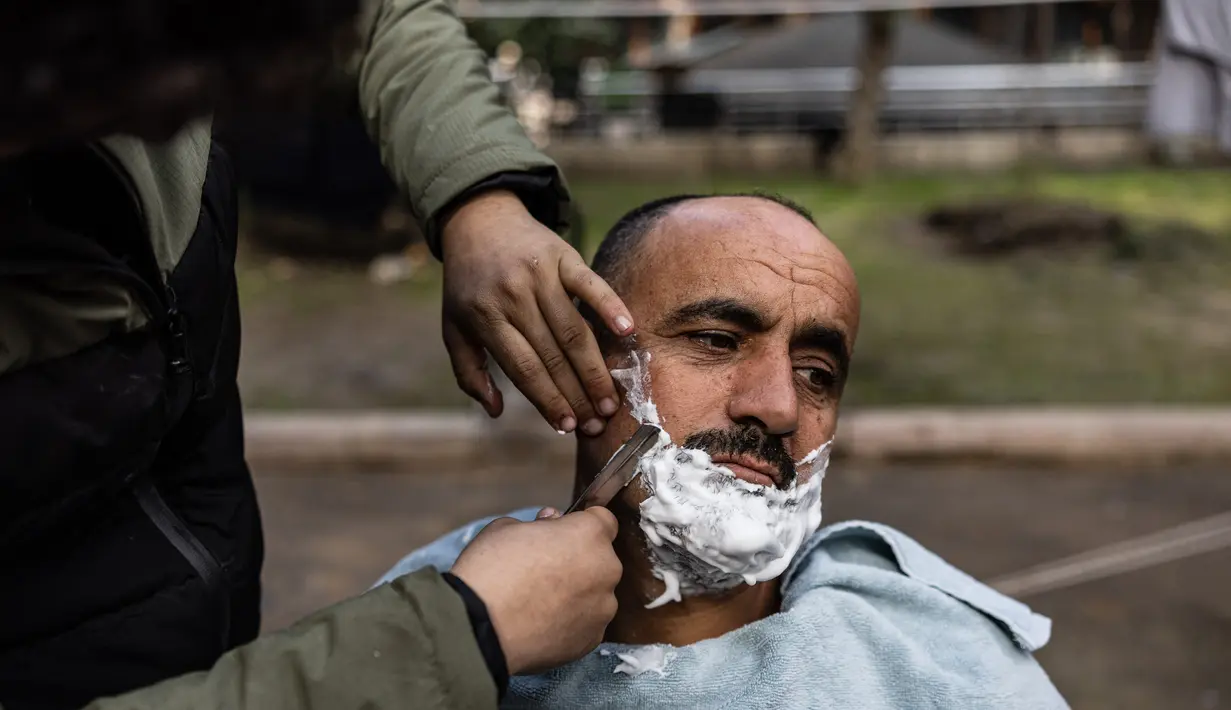 Khaled al-Hamo sedang dipotong rambut oleh putranya, Mohammed al-Hamo, di depan tenda mereka di kamp darurat di kota Antakya pada 19 Februari 2023. Hamo menawarkan jasa memotong rambut di ruang terbuka untuk keluarganya dan mereka yang kehilangan tempat tinggal akibat gempa dahsyat yang menimpa Turki dan Suriah pada 6 Februari 2023. (Sameer Al-DOUMY / AFP)