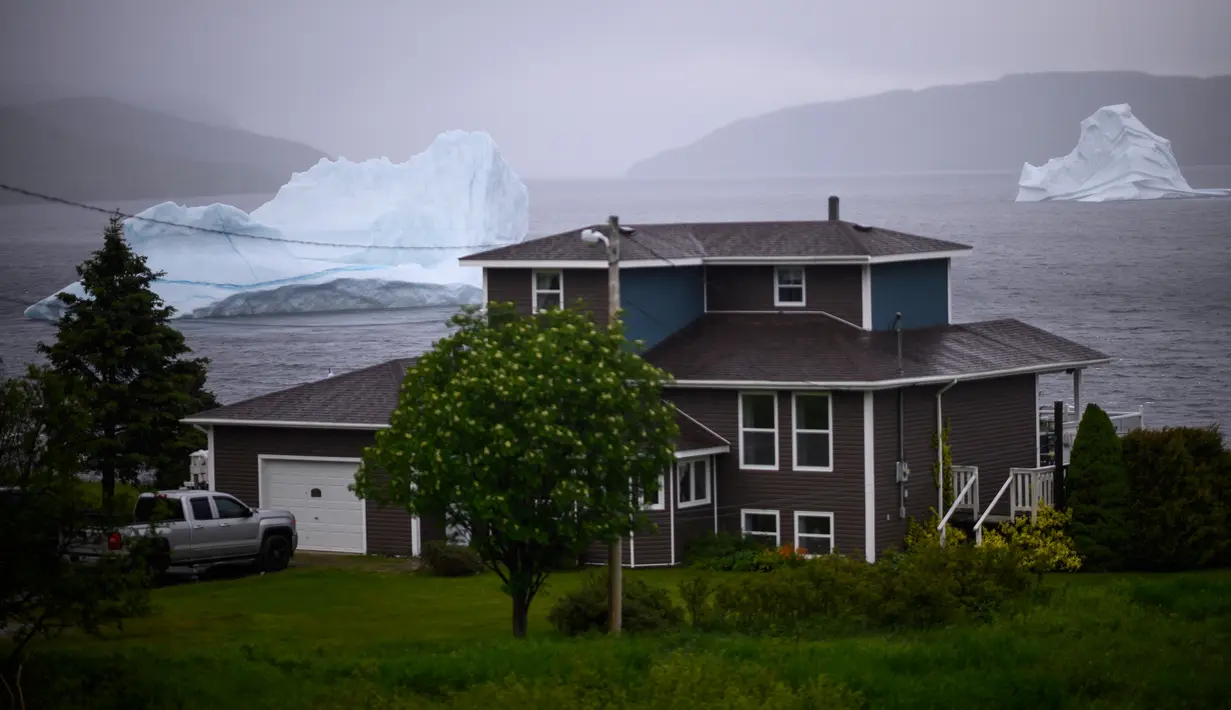 Gunung es terapung terlihat di balik rumah di King's Point, Newfoundland, Kanada, 2 Juli 2019. Potongan besar berwarna putih itu biasa mengapung sepanjang musim semi dan musim panas. (Johannes Eisele/AFP)