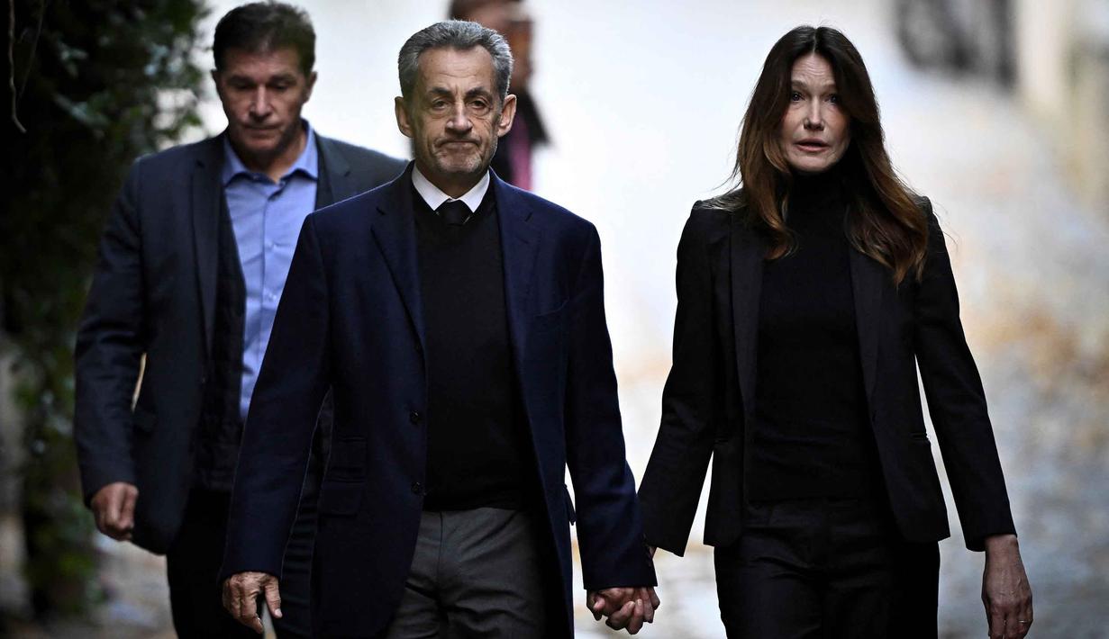 Mantan Presiden Prancis Nicolas Sarkozy (tengah), bersama istrinya Carla Bruni-Sarkozy, berjalan meninggalkan kediamannya untuk menyerahkan diri ke Penjara La Sante guna menjalani hukuman penjara lima tahun setelah dinyatakan bersalah atas konspirasi kriminal terkait rencana pendanaan kampanye pemilu 2007-nya oleh diktator Libya Moamer Kadhafi, di Paris, pada 21 Oktober 2025. (JULIEN DE ROSA/AFP)