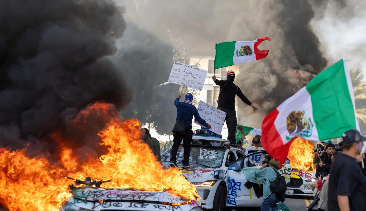 Pengunjuk rasa melambaikan bendera dari atas mobil yang rusak saat bentrok dengan penegak hukum di jalan-jalan di sekitar gedung federal selama protes setelah operasi imigrasi federal di Los Angeles, California, Amerika Serikat pada 8 Juni 2025. (RINGO CHIU/AFP)