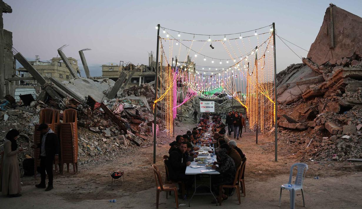 Di lokasi pengungsian, warga mencoba menghidupkan suasana Ramadan dengan kreativitas, seperti membuat lentera (fanous) dari kaleng bekas atau kain bantuan. Tampak dalam foto, warga Palestina duduk di meja panjang di tengah reruntuhan bangunan yang hancur untuk berbuka puasa bersama, selama bulan suci Ramadan di Khan Younis, Jalur Gaza, Kamis 19 Februari 2026. (AP Photo/Abdel Kareem Hana)