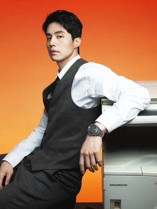 Mengenakan vest dan celana warna abu-abu, serta kemeja putih lengan panjang lengkap dengan dasinya, Lee Dong Wook mengenakan jam tangan strap stainless dengan dial hitam. [dok. Tissot]