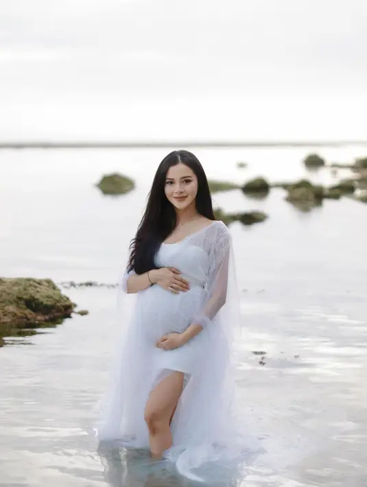 Senyum bahagia Bella Bonita menjalani maternity shoot di pinggir pantai di Bali. Bella tampak berpose dengan memegang perutnya yang makin besar. [Instagram/bellabonita_r.a]