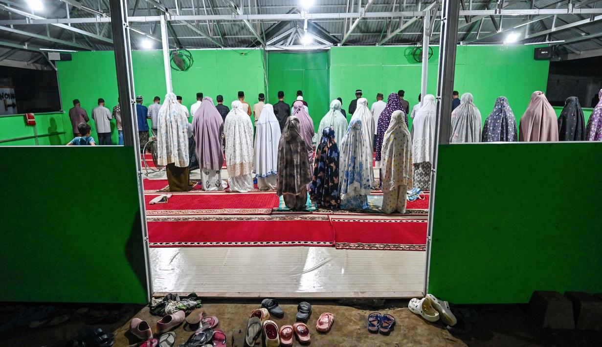 Diketahui, pemerintah setempat masih terus mengupayakan penyelesaian pembangunan Huntara. Tampak dalam foto, warga yang tinggal di tempat penampungan sementara bagi korban banjir melaksanakan salat berjamaah di sebuah aula di Meureudu, Kabupaten Pidie Jaya, Provinsi Aceh pada 21 Februari 2026, setelah banjir dan tanah longsor dahsyat melanda Sumatra, akhir November 2025 lalu. (CHAIDEER MAHYUDDIN/AFP)