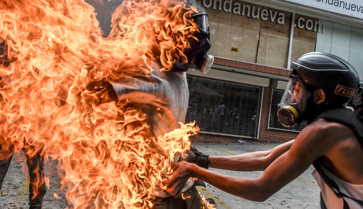 Demonstran melepaskan pakaian rekannya yang terbakar saat unjuk rasa yang diwarnai bentrokan menentang Presiden Venezuela, Nicolas Maduro di Caracas, Rabu (3/5). Pria itu tersulut api dari ledakan tangki motor milik polisi. (RONALDO SCHEMIDT / AFP)