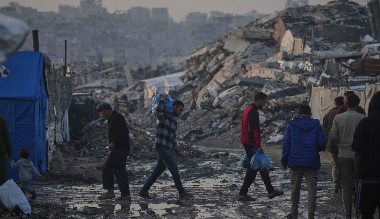 Fokus bantuan mencakup pengiriman pasokan medis ke fasilitas kesehatan yang tersisa serta peralatan untuk perbaikan layanan air dan sanitasi guna mencegah krisis penyakit. Tampak dalam foto, warga Palestina berjalan melewati perkemahan tenda di tengah bangunan-bangunan yang hancur akibat serangan udara dan darat Israel di Kota Gaza, Selasa 6 Januari 2026. (AP Photo/Jehad Alshrafi)