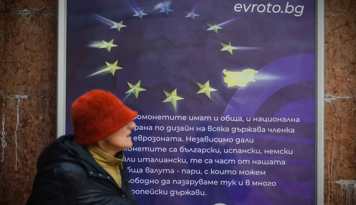 Mengadopsi euro dinilai sebagai fase akhir integrasi Bulgaria ke arus utama Eropa, setelah bergabung dengan NATO, Uni Eropa, kawasan Schengen, dan kini zona mata uang tunggal. Tampak dalam foto, seorang wanita berjalan melewati poster yang menampilkan Bulgaria sebagai anggota Zona Euro ke-21, di Sofia, Kamis 1 Januari 2026, saat negara tersebut bergabung dengan serikat mata uang euro. (AP Photo/Valentina Petrova)