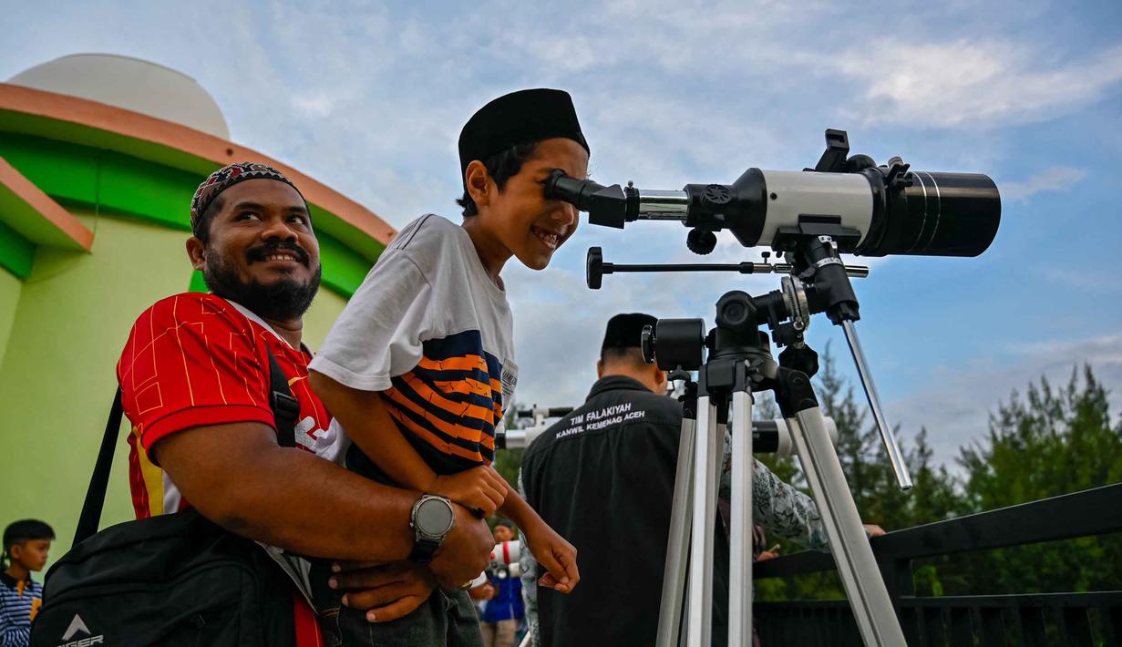 Untuk diketahui, metode rukyat sangat berbeda dengan hisab. Hisab murni menggunakan perhitungan matematis-astronomis tanpa memerlukan verifikasi visual di lapangan. Tampak dalam foto, seorang pria menggendong anaknya untuk mengamati bulan, menjelang Idul Fitri sekaligus menandai berakhirnya bulan suci Ramadan1447 Hijriah melalui teleskop di kantor observatorium, pantai Lhoknga, Aceh pada Kamis 19 Maret 2026. (CHAIDEER MAHYUDDIN/AFP)