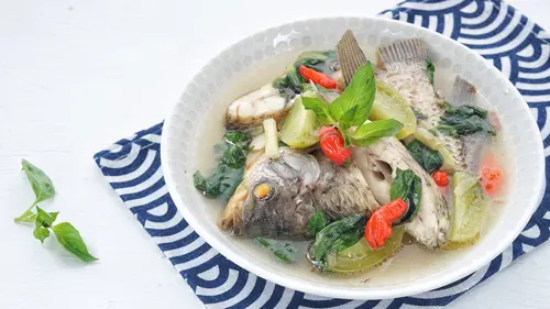 Semangkuk sup ikan nila hangat dengan kuah bening yang kaya rempah, dihiasi potongan wortel dan tomat segar