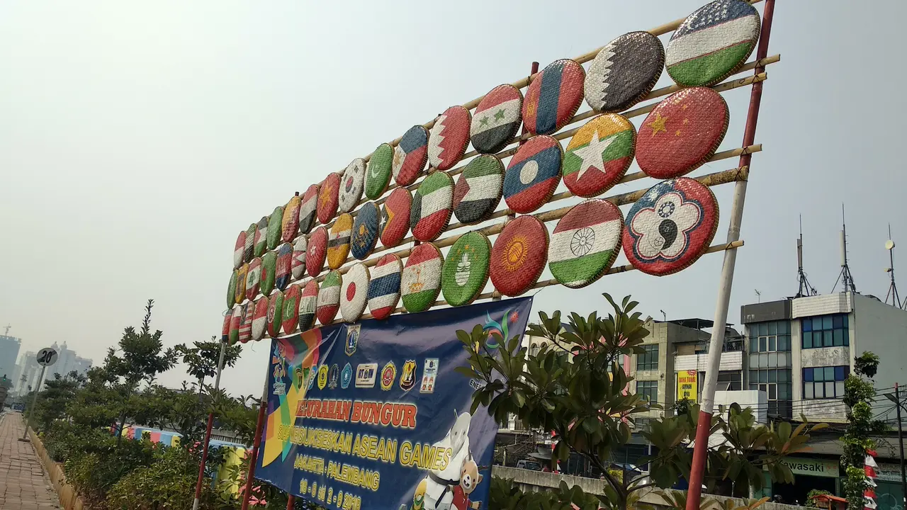 45 gambar bendera peserta Asian Games yang disajikan menggunakan tampah beras yang digantung pada tiang bambu di bantaran Kali Sentiong.  (Liputan6.com/Ika Defianti)